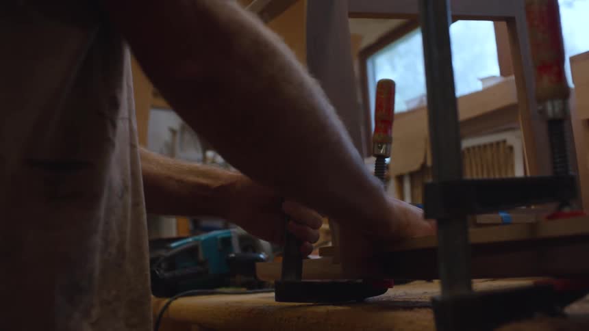 A person is working on a piece of wood using a tool in a cluttered workshop with various tools materials and a window in the background