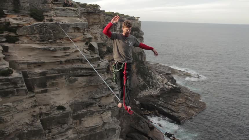 Man stands on tightrope between two cliffs before flipping around on a cloudy day