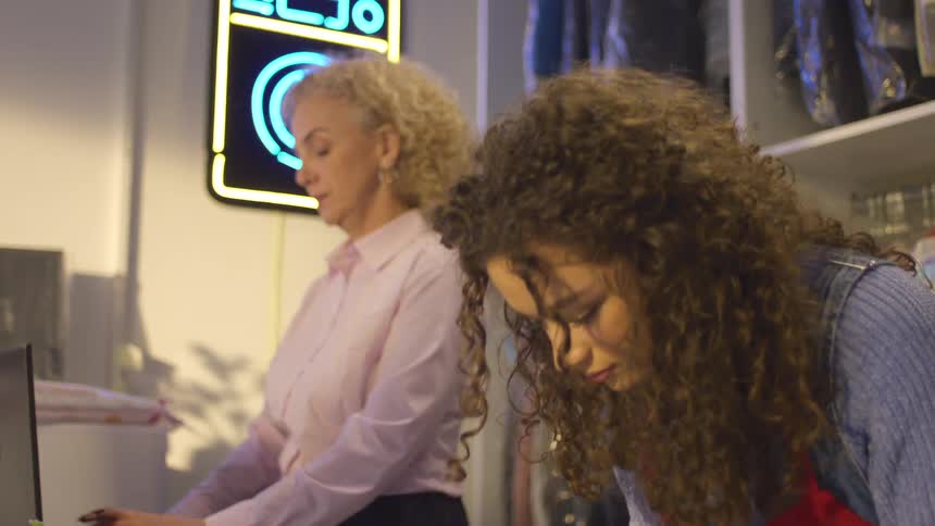 Curlyhaired woman taking notes on a notepad next to another woman typing on a laptop