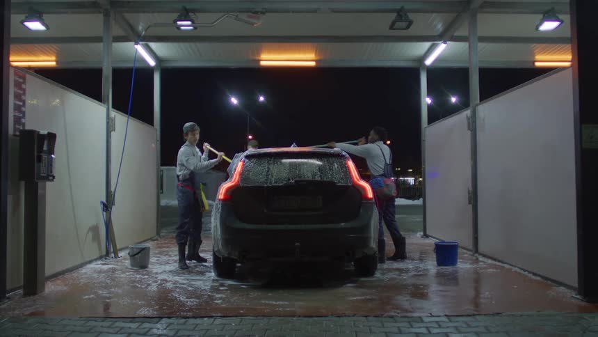 People are washing a car at a selfservice car wash at night with the car covered in soap and various cleaning supplies around them