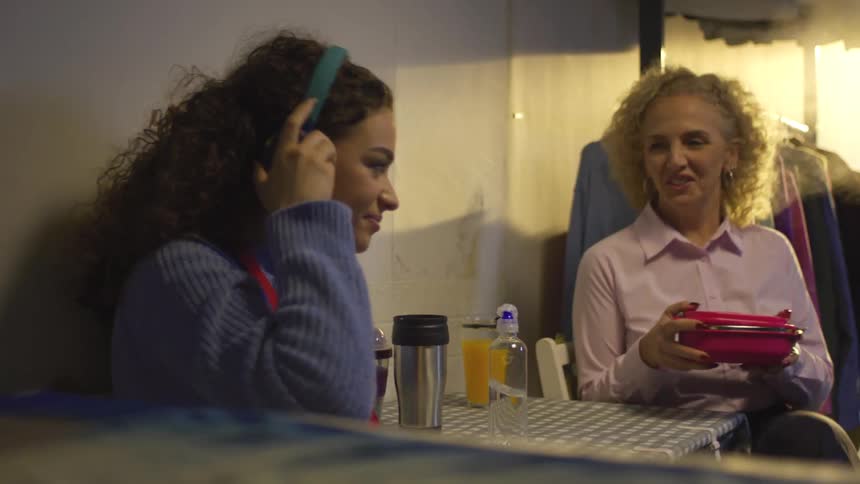 Woman sitting listening to music and taking off her headphones when another woman sits next to her and offers her a sandwich