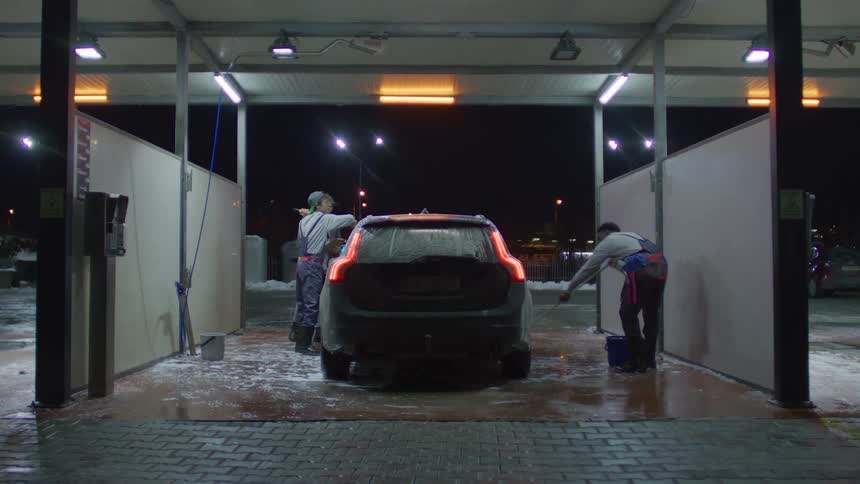 Three workers clean a car at night in a wash stall with two scrubbing the sides and another walking in front to observe while snow is visible in the background