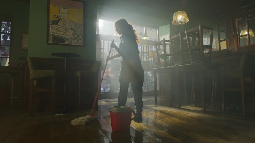 An employee is mopping the floor of a restaurant with chairs stacked on tables illuminated by sunlight streaming through a window