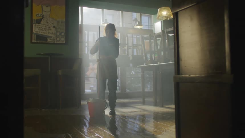 A woman is mopping the floor of a dimly lit restaurant with chairs stacked on tables illuminated by sunlight streaming through a window