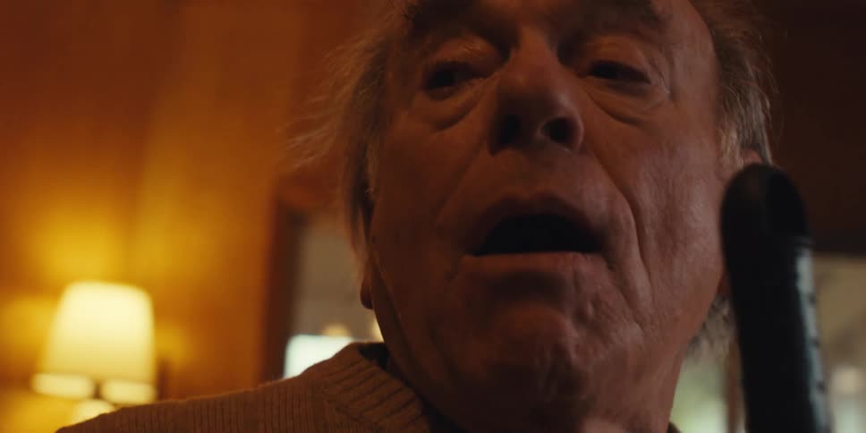 An elderly man is standing in a living room and he is now using a broomstick to sing