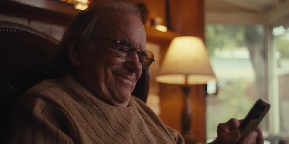 The face of an elderly man who is sitting on a sofa and is using his cell phone and starts to smile