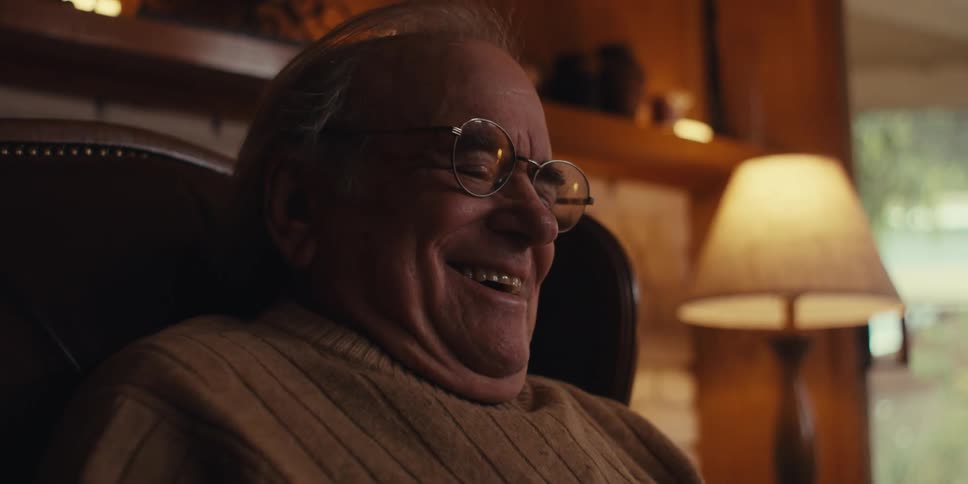 Elderly man with glasses wearing a beige sweater sitting in a dark leather armchair smiles while interacting with a smartphone