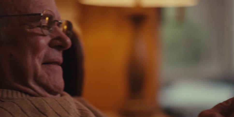 An elderly man is seated in a leather armchair in a cozy living room holding a smartphone