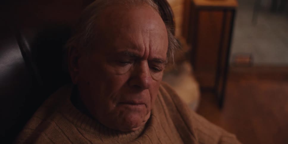 Elderly man wearing a beige sweater holds a smartphone while looking at the screen with a serious expression