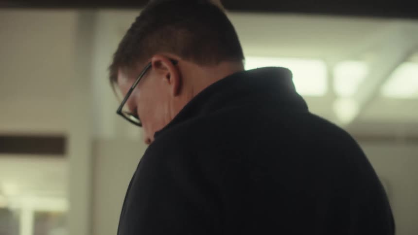 A middle aged man with glasses and a black turtleneck is captured in profile speaking on a smartphone with a serious expression in a softly lit indoor setting