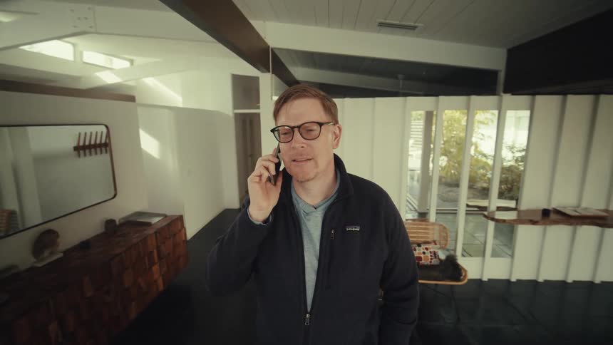 A middle aged man with glasses and a dark jacket is standing in a modern home holding a smartphone to his ear while engaged in a conversation looking out the window with a focused expression