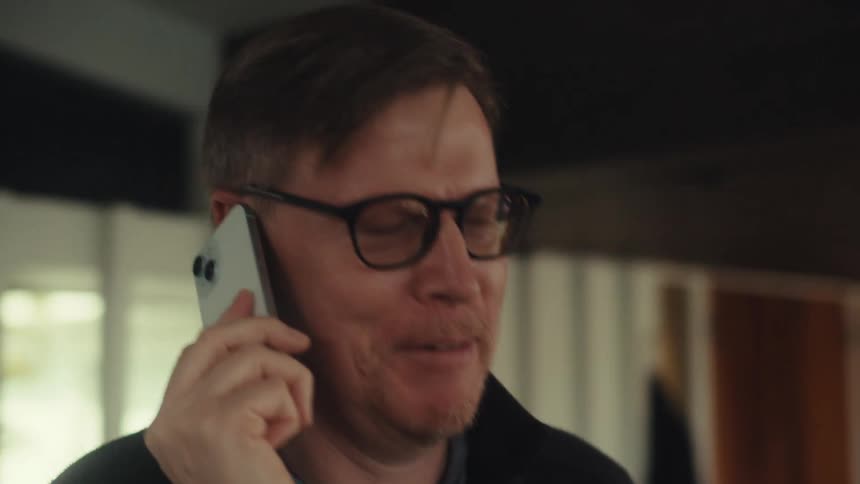 A middle aged man with glasses and a dark jacket talks on his smartphone nodding his head in agreement