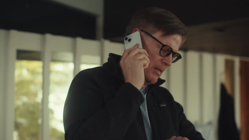 A middle aged man with glasses and a dark jacket talks on his smartphone checking his watch