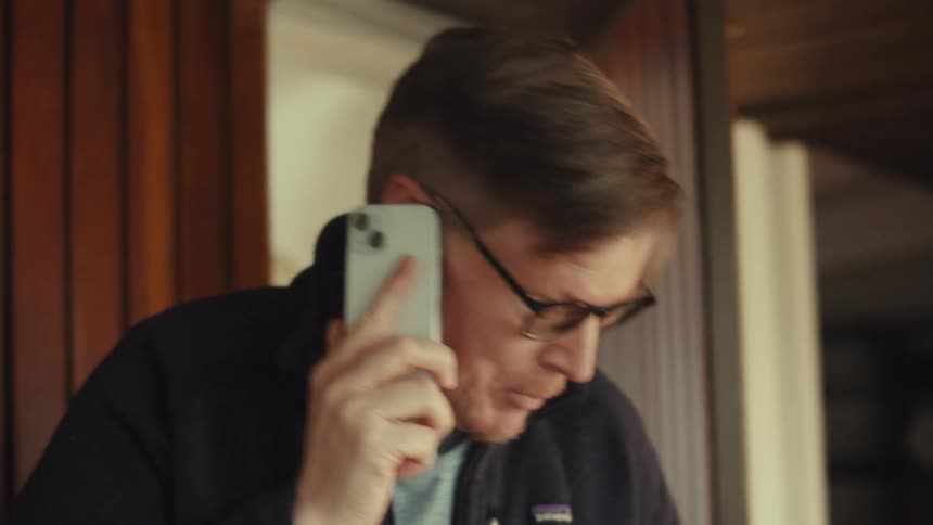 A middle aged man with glasses and a dark jacket talks on his smartphone while opening the door with a bag in his hand