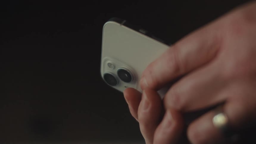 A hand holding a silver iphone highlighting its dual rear camera system against a blurred background