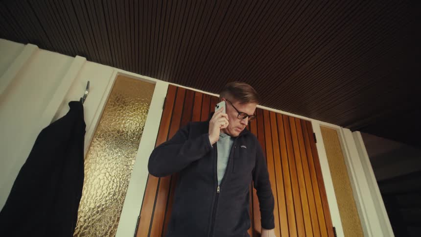 Middle aged man wearing glasses and a black jacket talks on the smartphone while opening the wooden door