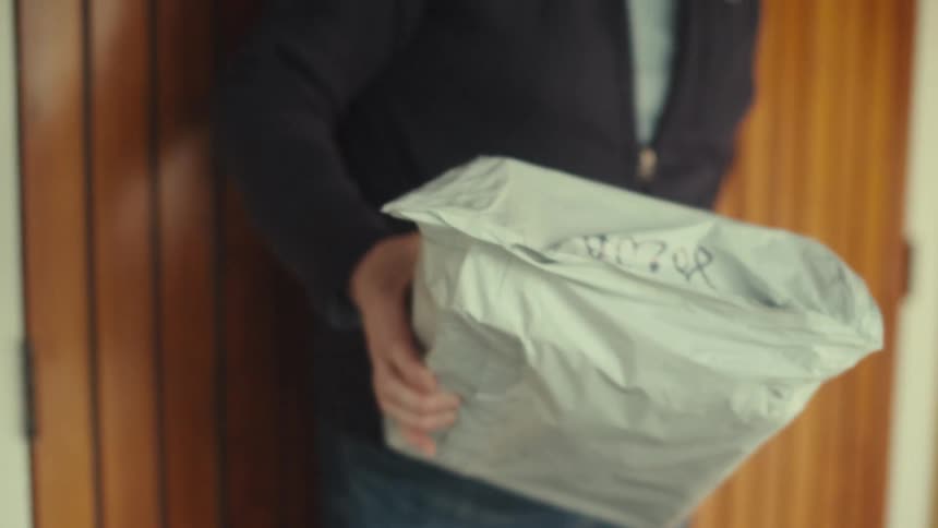 A man wearing glasses and a black jacket opens a wooden door and enters a room holding a crumpled light colored package in front of him