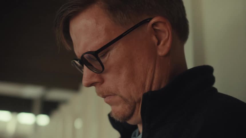 A middle aged man in a dark jacket and glasses uses a silver iphone near the door