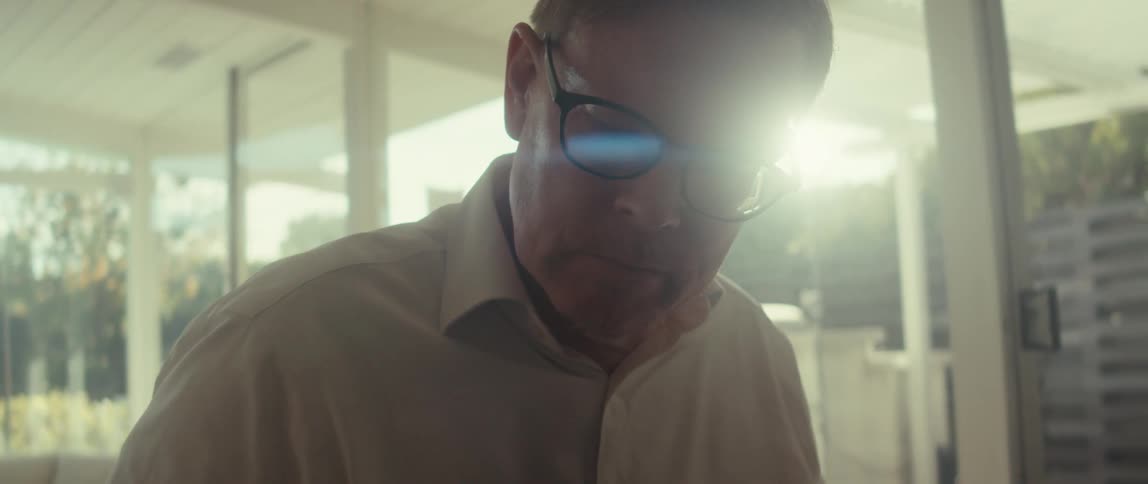 The face of an adult man wearing glasses who is looking at something in front of him and nodding in confirmation and behind him a wall of glass where sunlight is passing through