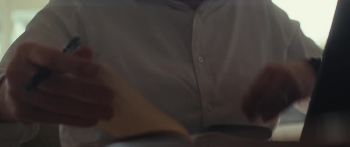 Man in a white shirt and a watch on his wrist flips through a notebook while holding a pen next to a laptop