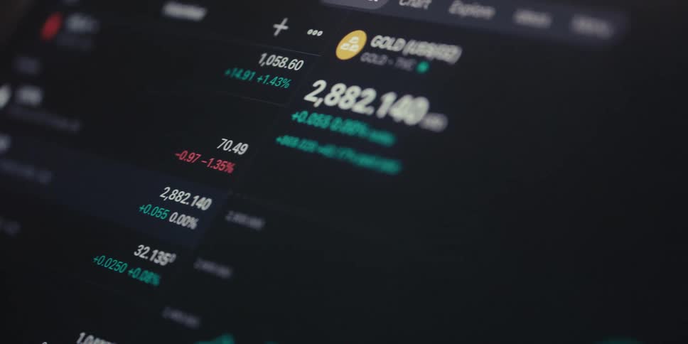 An ipad screen displays various currency and crypto prices highlighting gold at 2882140 as a hand scrolls with an apple pencil