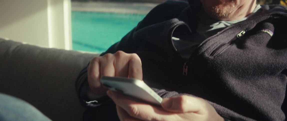 A man with glasses wearing a black jacket and jeans sits on the couch in the living room using a silver iphone near the swimming pool