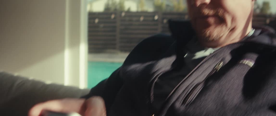 A man with sunglasses and a black jacket sits on the couch in the living room looking down at his smartphone a swimming pool is visible in the background