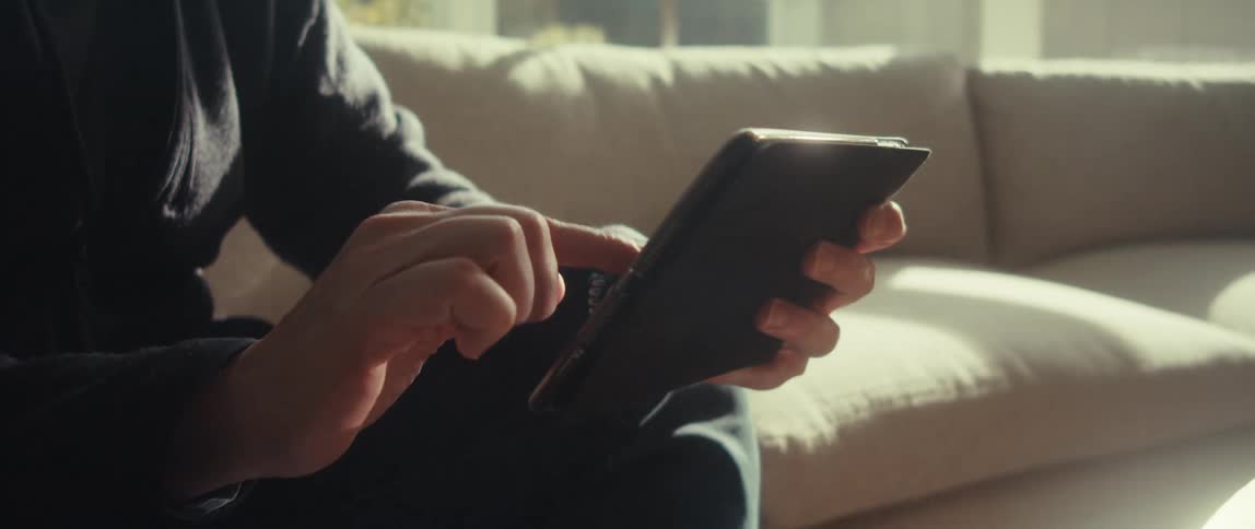 A man is shown holding a tablet in their hands with their torso and part of a couch visible in the background