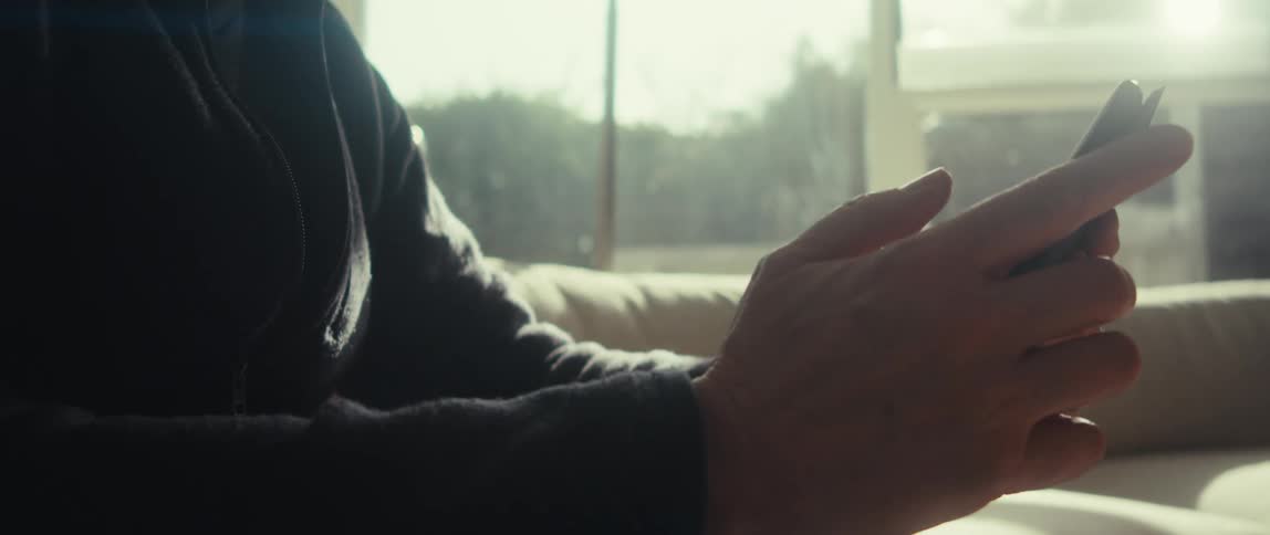 A person is sitting on a couch in a sunlit room holding and using a smartphone with both hands