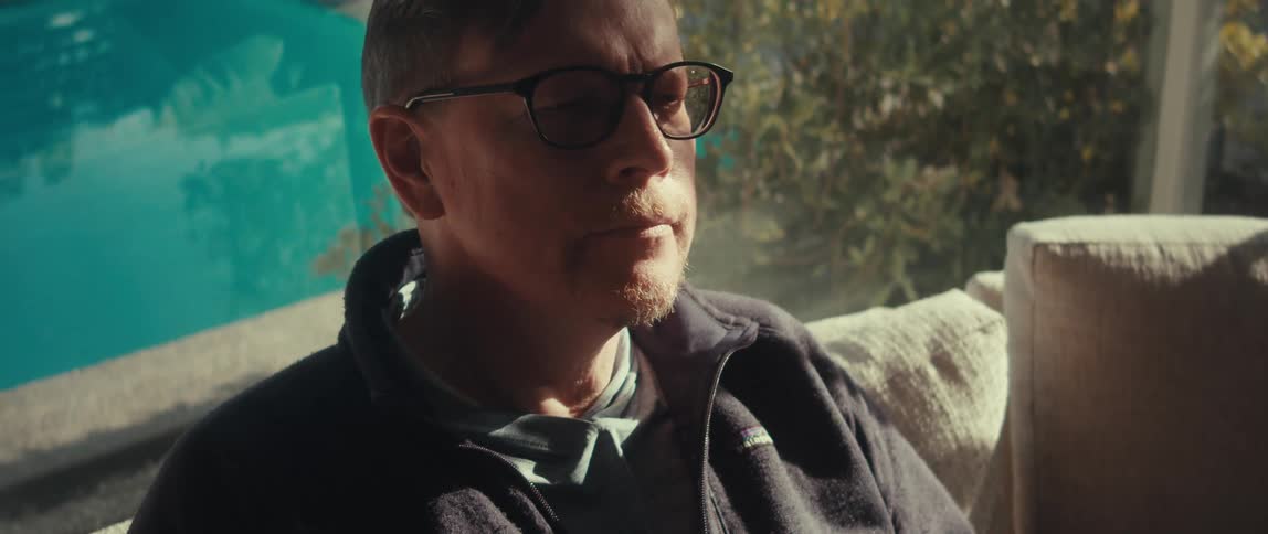 A middleaged man wearing sunglasses and a black jacket sits on the couch in the living room using a smartphone next to the swimming pool
