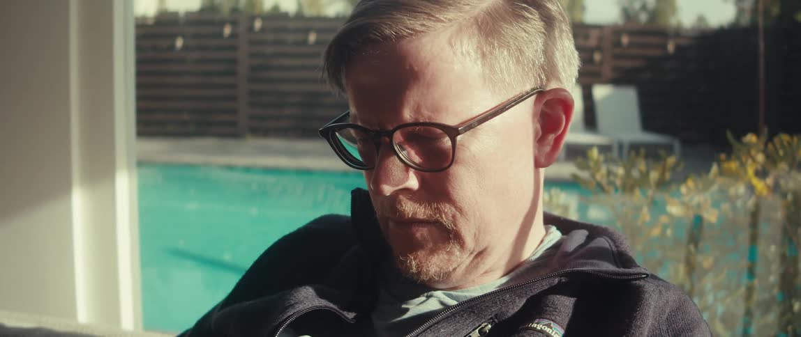 A man with glasses and a goatee looks down thoughtfully near a pool