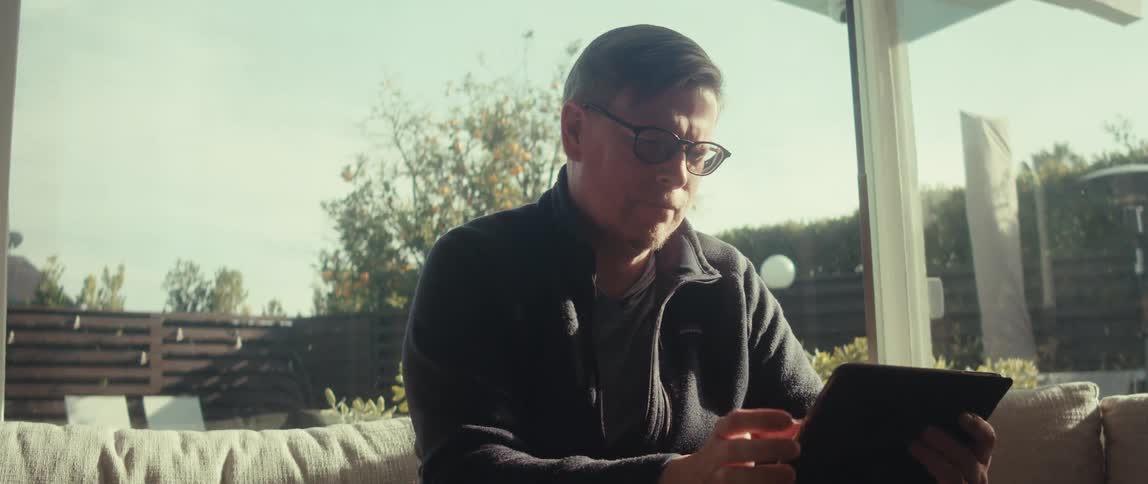 An older man with glasses in a grey shirt and dark hoodie sits on a sofa in an enclosed patio with large windows scrolling on a tablet