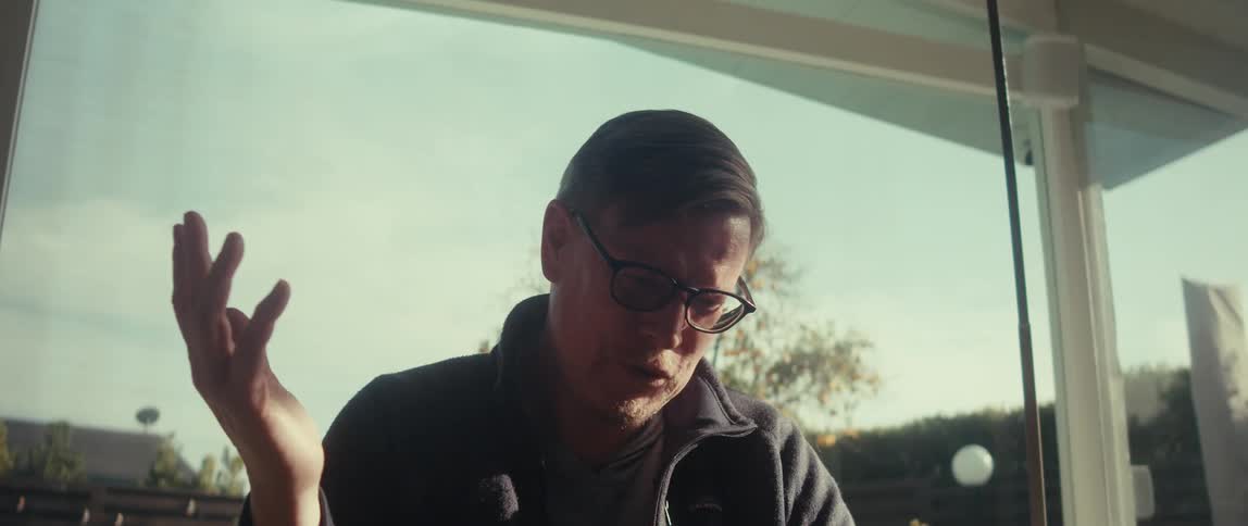 A man with glasses gestures and talks in front of a window