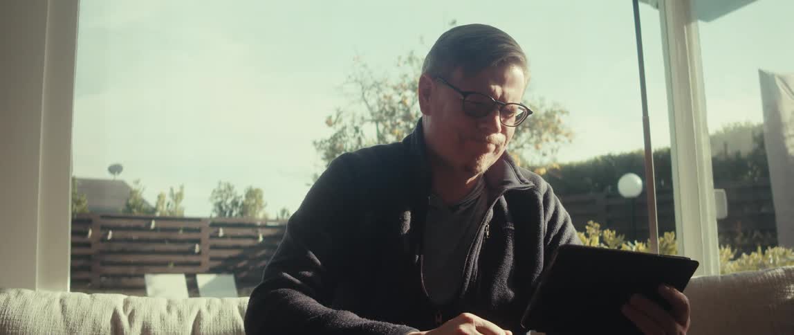 Man in glasses and a dark jacket uses a tablet while sitting on a sunlit sofa