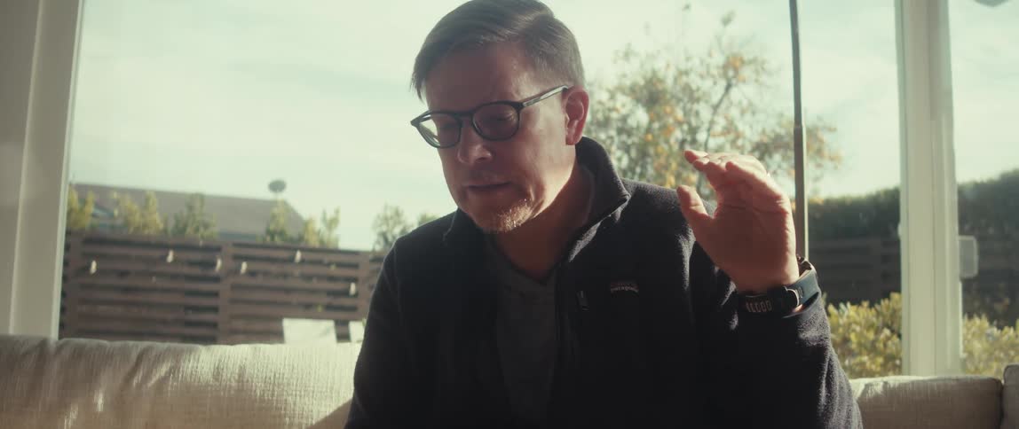 A man sitting on a couch in a brightly lit sunny room is explaining something while gesturing