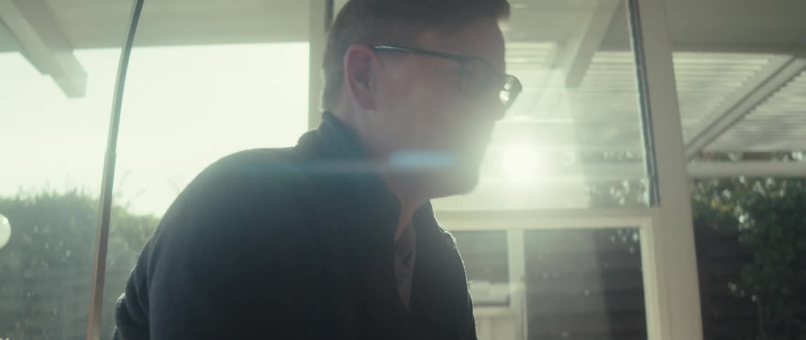An older man sits in an enclosed patio appearing to converse with someone offscreen