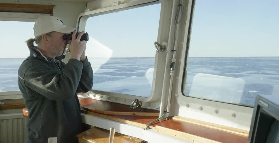 Ponytailed man looking out at the cold water from inside his ship using his binoculars