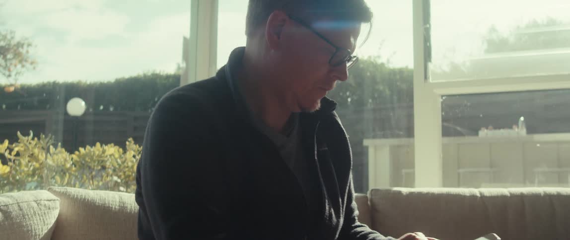 A middle aged man wearing glasses and a dark sweater sits on a couch near a large window looking at his smartphone with a focused expression
