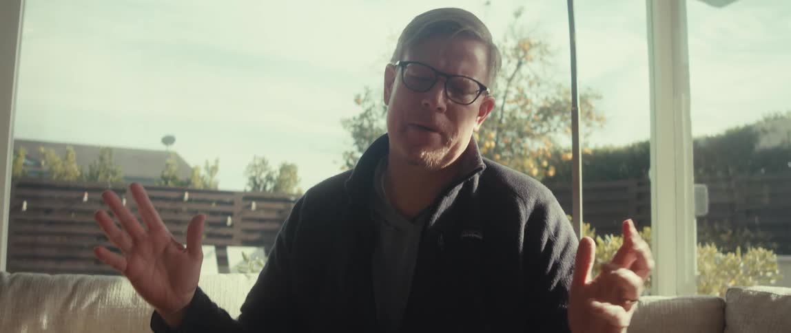 A middle aged man with glasses and a black jacket is on a video call on his laptop in the living room