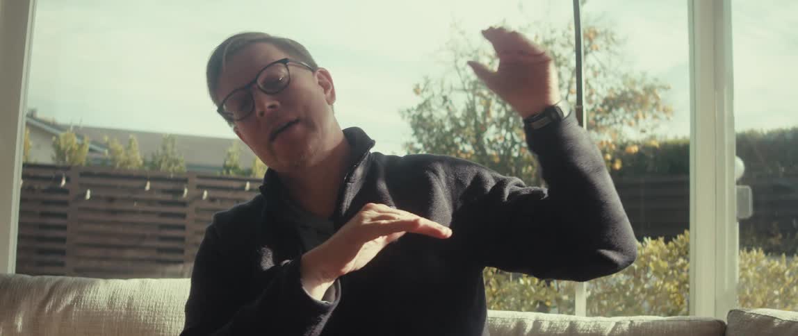 A man in glasses sitting on a couch gestures with his hands while talking to someone in a bright room
