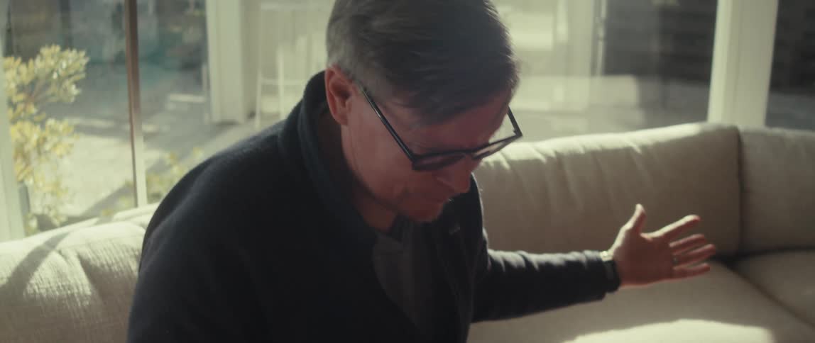 An older man uses hand gestures while speaking to someone offscreen seated on a couch in an enclosed patio with large windows