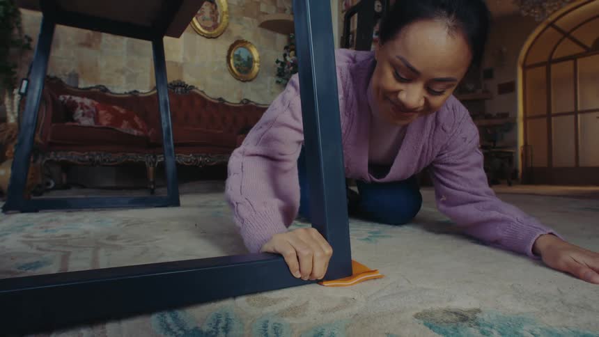 A person is kneeling on a carpeted floor adjusting the leg of a table by placing an orange wedge under it with a vintage sofa and framed pictures visible in the background
