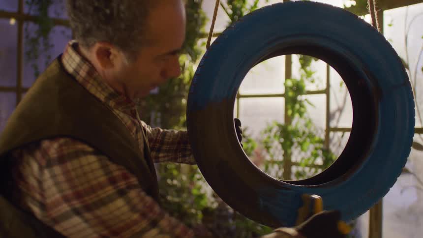 Man painting a tire blue while holding a rope in a greenhouse