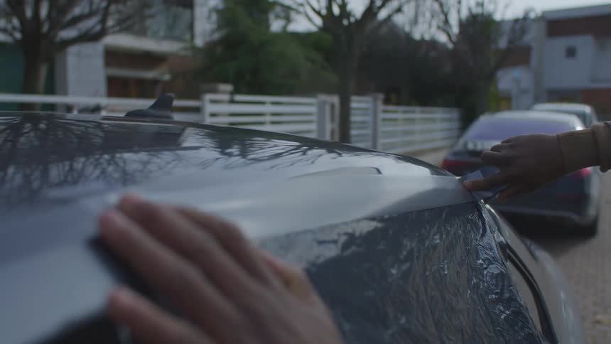 A person is applying a strip of duct tape to a car window in a tranquil neighborhood in the morning