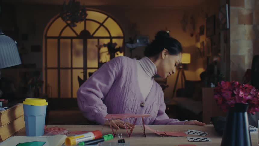 A woman in a purple sweater is building a house of cards on a table that is crumbling in a cozy room