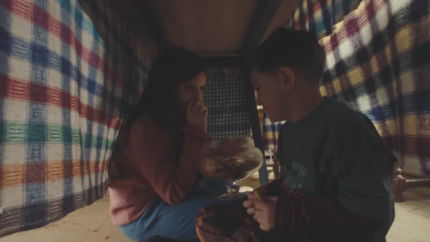 Brother and sister are hiding under a table with a tablecloth eating cookies from a jar