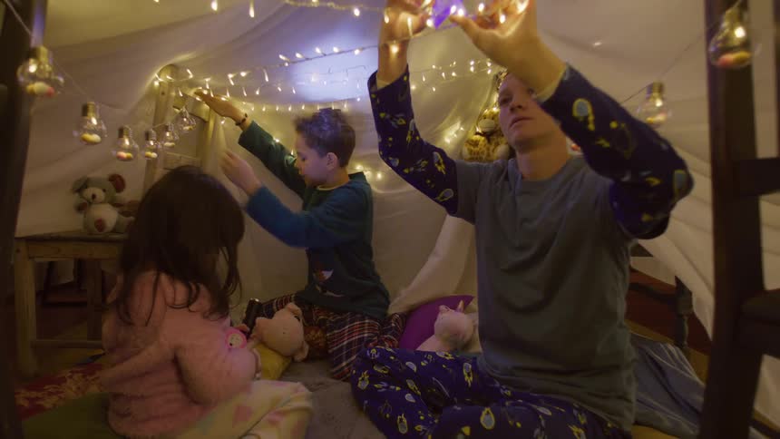 Three small children playing in a hut made by a sheet and decorated with lights