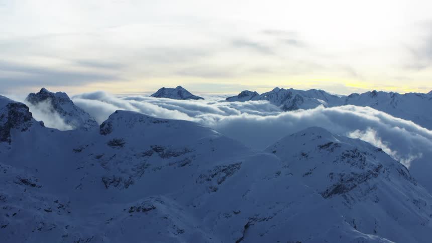 Snowcovered mountain range with dense clouds between the peaks and a sky partially lit by the setting sun