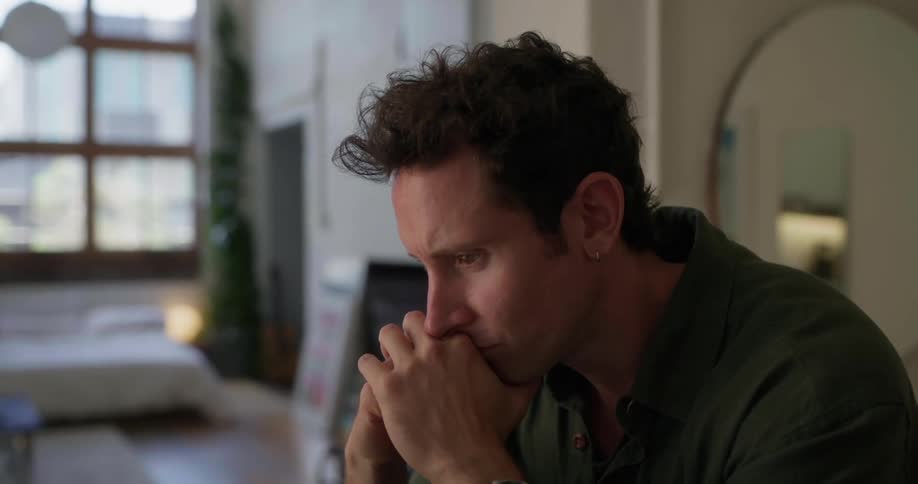 A man sitting in a green shirt is looking forward with his hands on his chin thoughtfully and then gives a smile