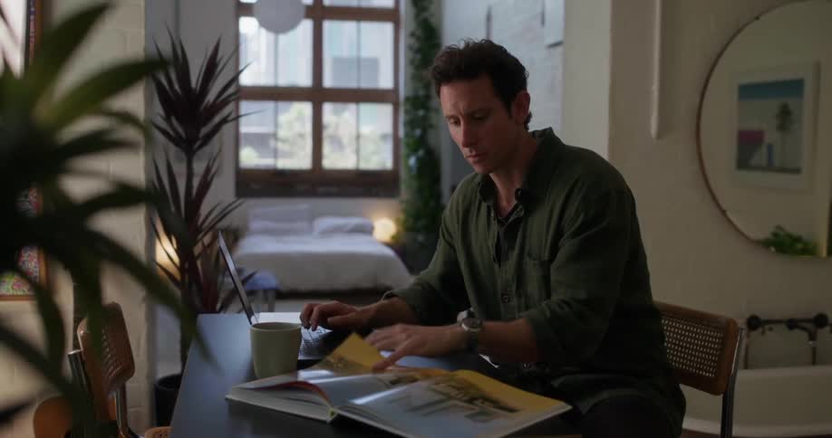 A person is sitting at a table in a welllit room with large windows flipping through a magazine with a laptop and a mug on the table and a bed visible in the background